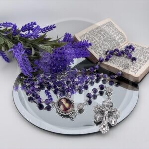 A purple catholic rosary with a photograph of St. Mary Magdalene in the centre and a silver crucifix laying on a mirror close to a purple plant and a prayer book